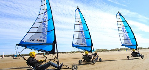 Land yachts with blue sails racing across the sandy beach under a bright blue sky.
