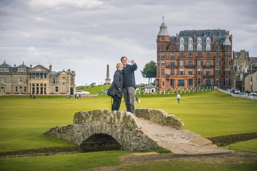 Two people taking a selfie on the Swilcan Bridge at St Andrews Old Course with historic buildings behind.