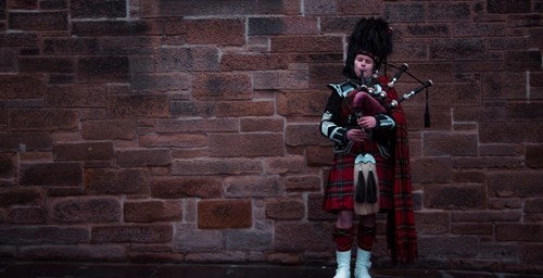 Bagpiper in full traditional Scottish attire playing in front of a red sandstone wall.