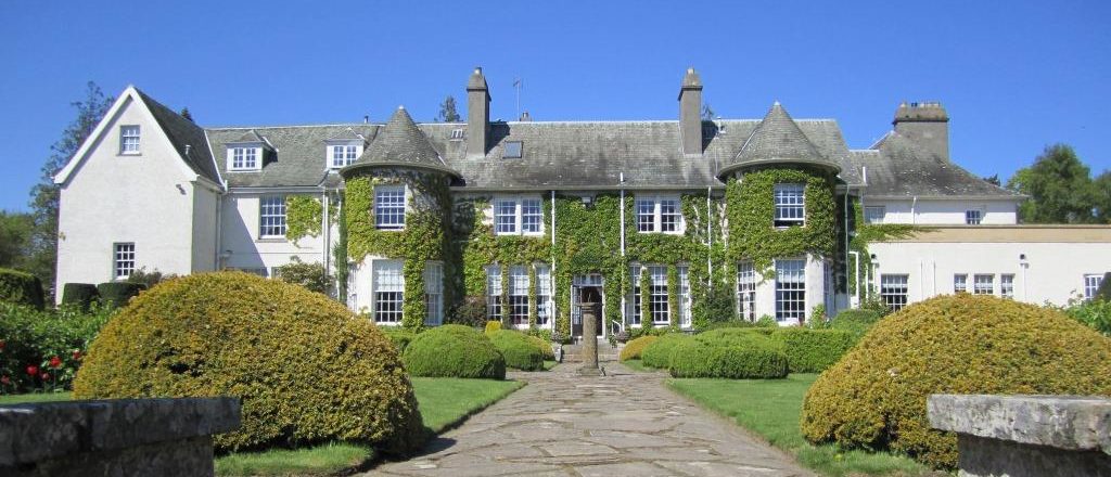 The grand historic manor house of Rufflets St Andrews hotel with ivy-covered walls, garden and trimmed bushes under a clear blue sky.