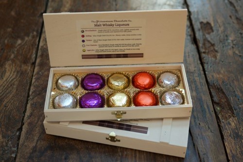 Assorted malt whisky liqueur chocolates in colorful foil inside a cream gift box on a wooden table.