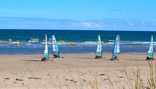 Land yachts with blue sails racing along a sandy beach with kayakers paddling in the sea.