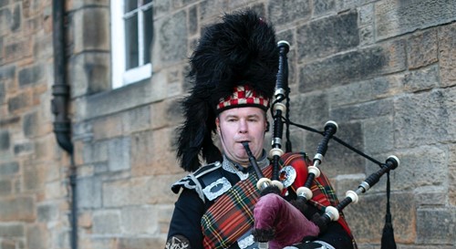 Bagpiper in traditional Scottish attire playing outside a stone building.