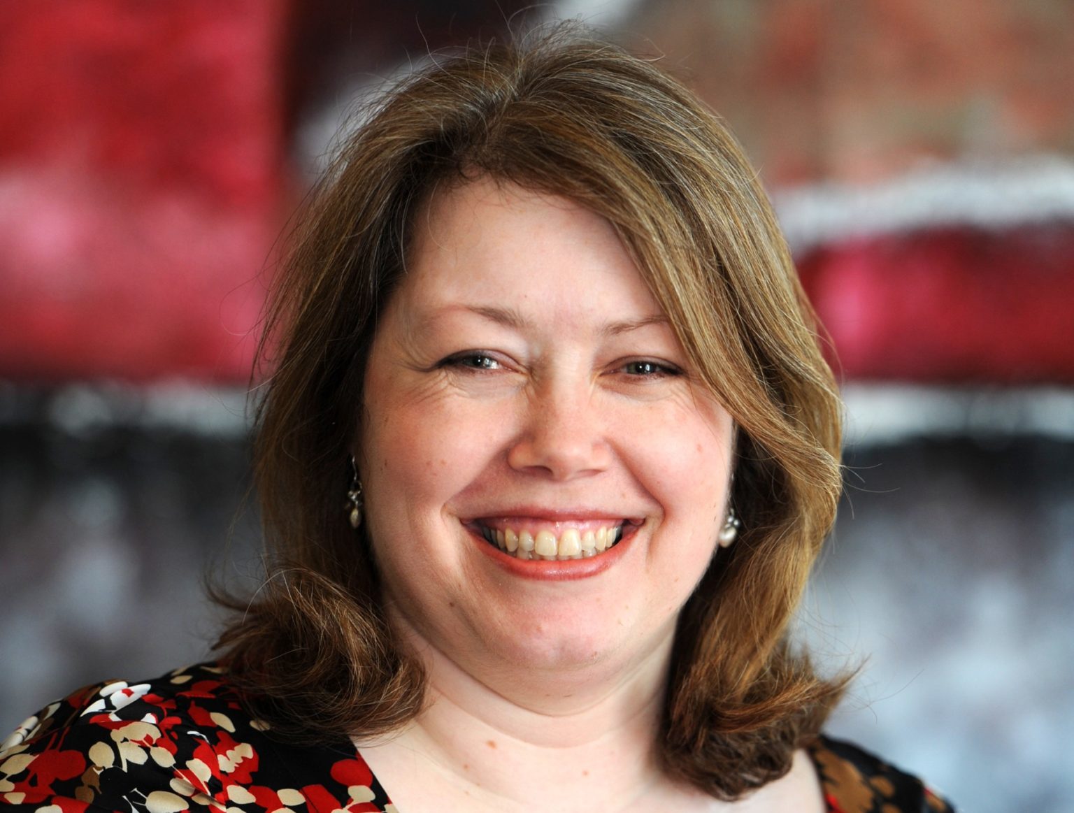 Close-up of a woman with shoulder-length hair smiling in front of a blurred red and black background.