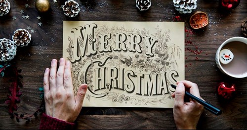 Hands drawing “Merry Christmas” in vintage lettering surrounded by festive items and a hot drink.