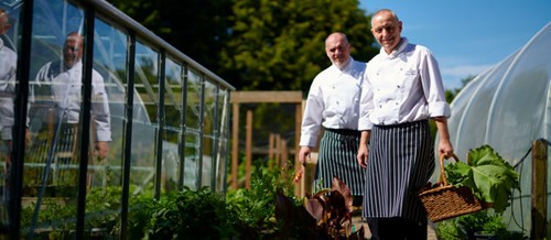 Two chefs walking through a garden beside a greenhouse, one holding a basket of fresh produce.