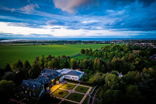 Aerial view of Rufflets St Andrews surrounded by green fields and dramatic clouds over the coastline.