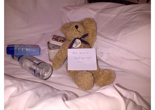 Teddy bear on white bedding with snacks, water and a note reading “It’s been a hard day’s night.”